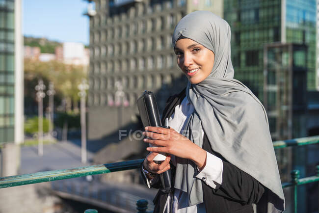 Empreendedora muçulmana alegre no hijab e com café takeaway em pé na rua olhando para a câmera — Fotografia de Stock