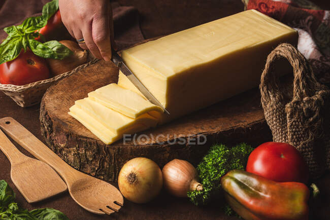 Blocco di formaggio su supporto di legno vicino a cipolle crude e sacchetto intrecciato contro spatole biologiche e foglie di basilico — Foto stock