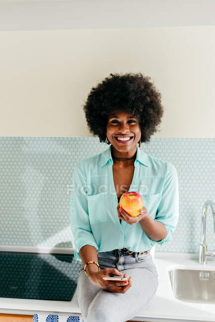 Moderna mulher afro-americana bonita com smartphone na mão sentado no balcão da cozinha olhando para a câmera sorrindo em casa e comendo maçã — Fotografia de Stock