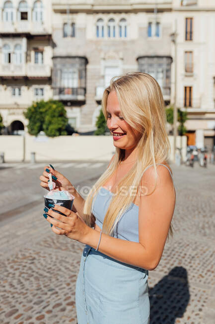 Beautiful blonde young female eating cold tasty ice cream while standing in city street on sunny day in summer — Stock Photo
