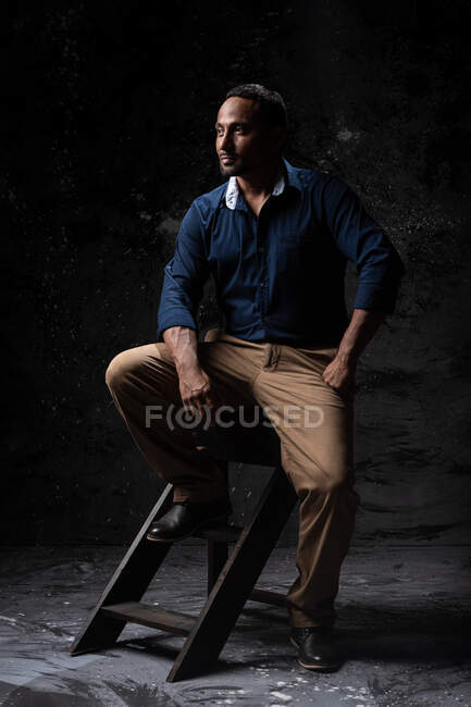 Серьезный стильный этнический мужчина сидит на деревянном стуле в студии на темном фоне и смотрит в сторону — стоковое фото