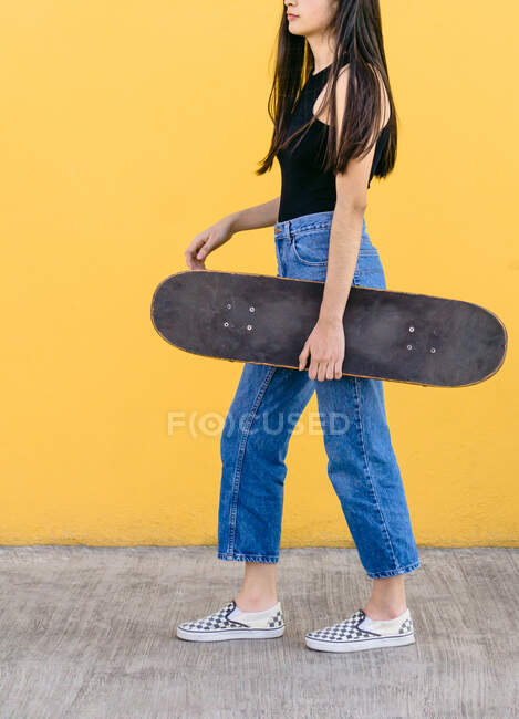 Cortado irreconocible joven patinadora femenina con monopatín de pie mirando hacia otro lado en la pasarela con colorida pared amarilla en el fondo durante el día - foto de stock