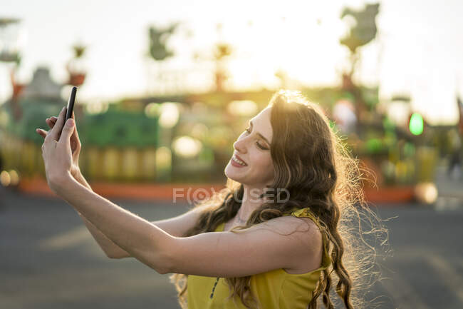 Зміст жінки, яка приймає власний портрет на мобільному телефоні, розважаючись в парку розваг ввечері влітку — стокове фото