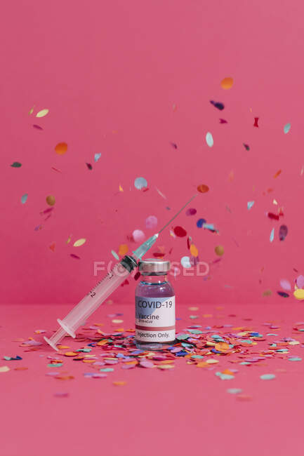 Frasco de vacuna contra el Coronavirus cerca de la jeringa con aguja sobre fondo rosa cubierto de confeti - foto de stock