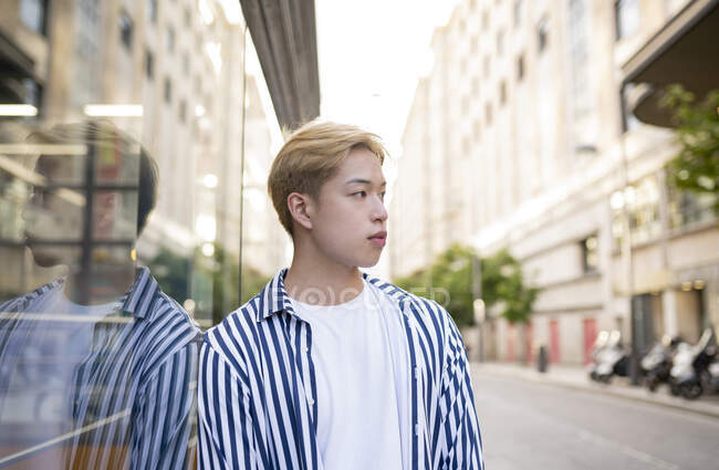 Stilvolles asiatisches männliches Modell steht in der Nähe von Gebäuden und reflektiert in einer verspiegelten Glaswand — Stockfoto