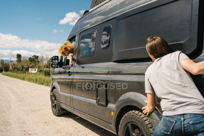 Vista trasera de un hombre joven empujando furgoneta mientras que la mujer sentada en el volante como teniendo problemas durante el viaje en el campo de verano - foto de stock