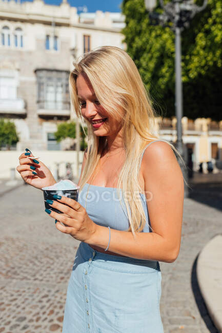 Hermosa rubia joven hembra comiendo helado frío sabroso mientras está de pie en la calle de la ciudad en el día soleado en verano - foto de stock