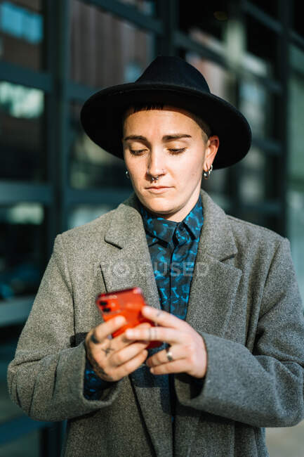 Androgyne personne en chapeau naviguant sur un téléphone portable en regardant l'écran debout dans la rue en plein jour — Photo de stock