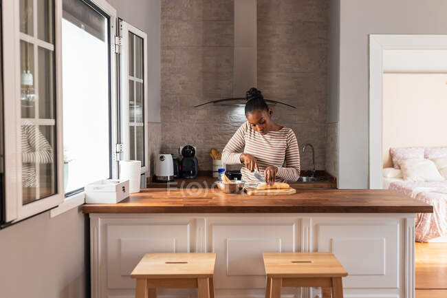 Mulher preta em vestuário com ornamento listrado cortando banana cozinhando com faca na tábua de corte na mesa em casa — Fotografia de Stock