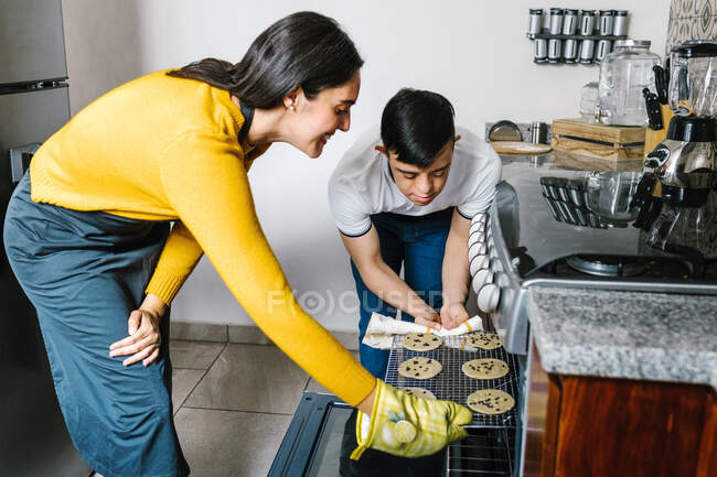 Mère ethnique heureuse dans tablier et adolescent avec trisomie 21 mettre des biscuits aux pépites de chocolat cru au four pour la cuisson dans la cuisine — Photo de stock