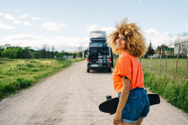 Felice donna afroamericana che porta il longboard e distoglie lo sguardo con il sorriso mentre cammina sulla strada di campagna vicino al furgone in estate — Foto stock