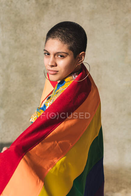 Серьезные молодые бисексуальные этнические женщины с разноцветным флагом, представляющим символы ЛГБТК и смотрящие в камеру в солнечный день — стоковое фото