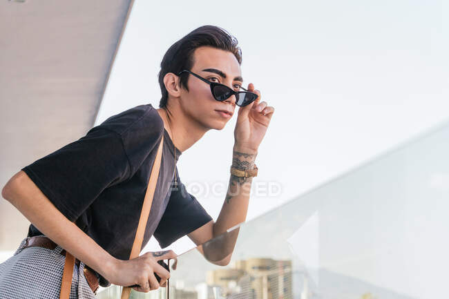 Basse angle vue latérale de gay mâle dans les lunettes de soleil à la mode penché sur balustrade en verre sur balcon et regardant loin — Photo de stock