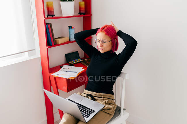 Очаровательная девушка-фрилансер в повседневной одежде, сидящая на стуле и использующая ноутбук во время работы над проектом в светлой современной квартире — стоковое фото