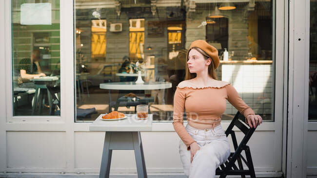 Trendige Französin in Baskenmütze sitzt am Tisch mit leckerem Kaffee und süßem Croissant beim Frühstück im Straßencafé — Stockfoto
