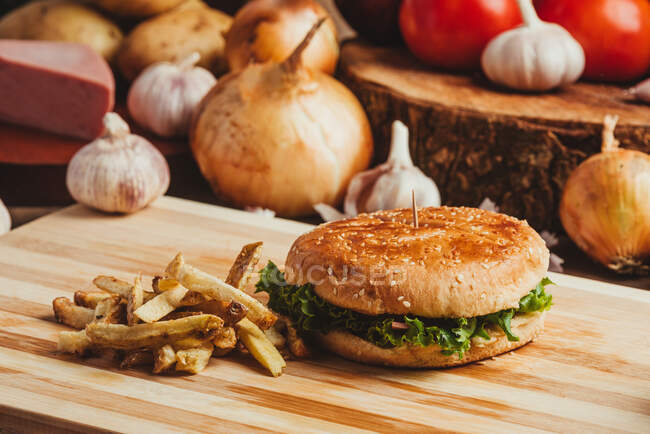 Hamburguesas apetitosas con verduras colocadas sobre tabla de madera con papas fritas en la cocina - foto de stock
