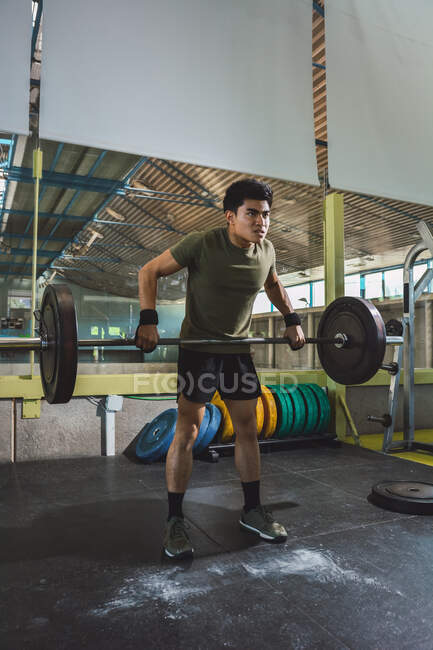 Focalisé asiatique athlète masculin faisant haltérophilie avec haltère lourd pendant l'entraînement dans la salle de gym détournant les yeux — Photo de stock