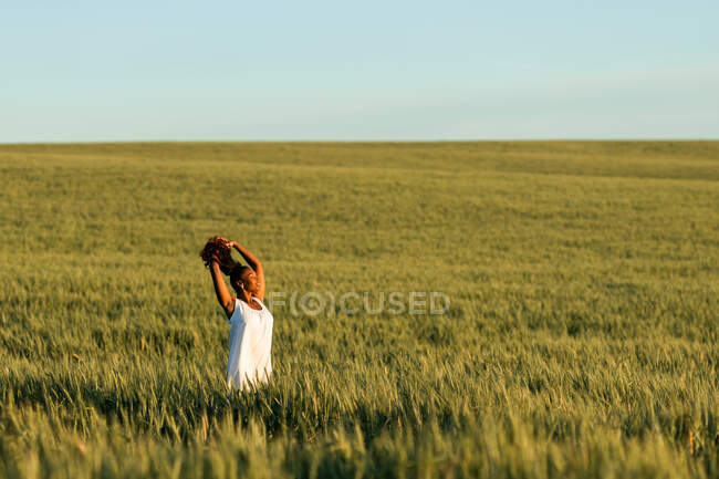 Young black lady in white summer dress strolling on green wheat field while touching hair in daytime under blue sky — Stock Photo