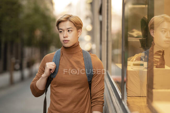Modelo masculino étnico seguro con cabello rubio y cuello alto de moda parado en la ciudad y mirando a la cámara - foto de stock