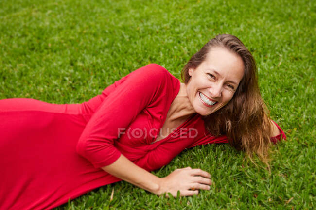 Жінка, одягнена в червоний, лежить на землі в парку з травою і дивиться на камеру — стокове фото