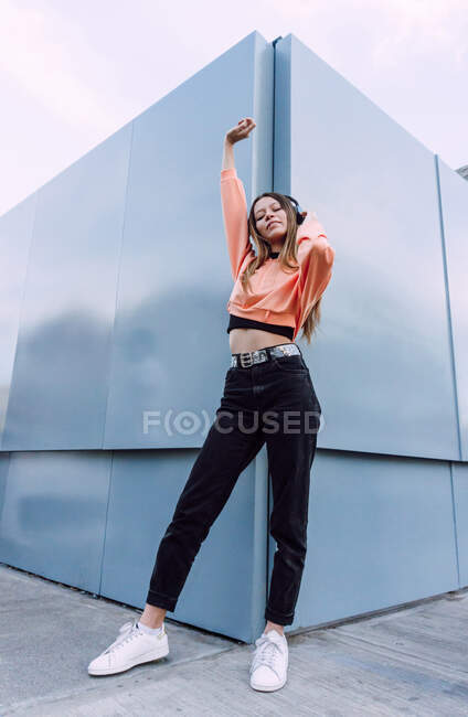 Низкий угол всего тела стильной женщины, стоящей возле современного геометрического здания и слушающей музыку в беспроводных наушниках — стоковое фото