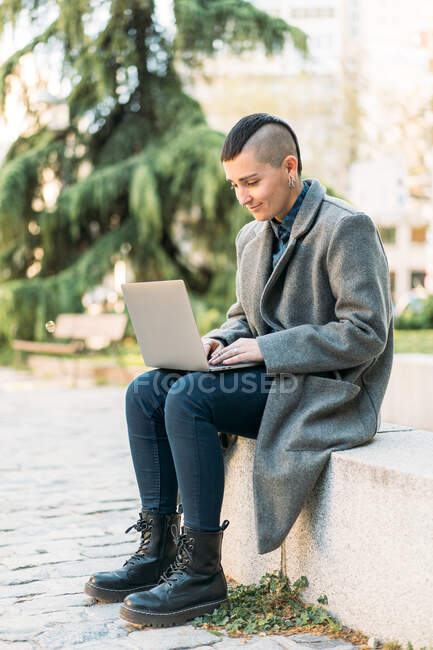 Androgynous persona con mohawk en botas y abrigo navegar por Internet en netbook mientras está sentado en la ciudad - foto de stock