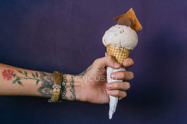 Mão tatuada de corte de macho com sorvete doce em cone de waffle no fundo roxo — Fotografia de Stock