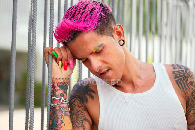 Queer male with bright pink hair and colorful nails standing in street and leaning on metal fence while looking down — Stock Photo