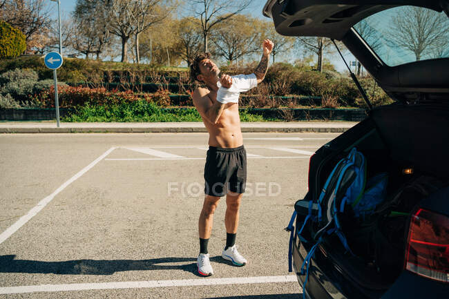 Masculine male athlete with tattoo putting on t shirt against automobile in parking on sunny day — Stock Photo