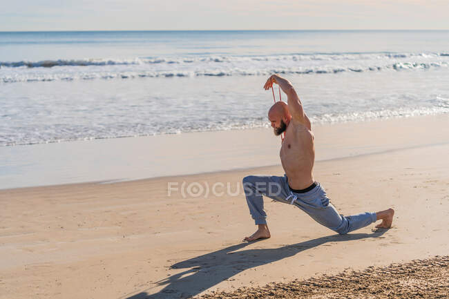 Side view of shirtless male athlete stretching arms with elastic band while doing a lounge working out on empty sunny beach — Stock Photo