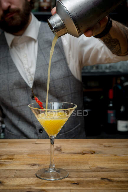 Cosecha irreconocible barman verter alcohol cóctel naranja de coctelera en vidrio colocado en el mostrador de madera en el bar - foto de stock