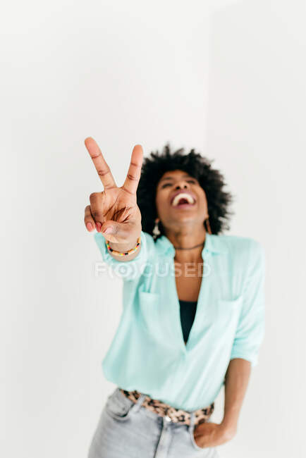 Playful young African American female in trendy outfit having fun and showing peace sign on white background — Stock Photo