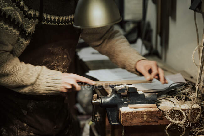 Vista laterale dell'anonimo artigiano maschio ritagliato usando la pietra abrasiva mentre levigava il piccolo dettaglio in legno della chitarra al banco da lavoro — Foto stock