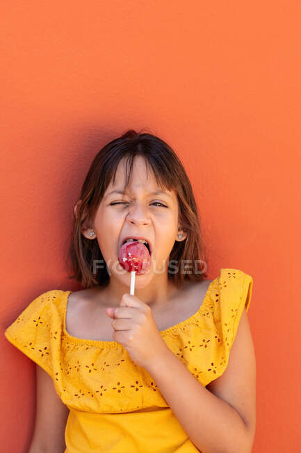 Child in yellow wear with floral ornament making face while enjoying tasty lolly and looking at camera — Stock Photo