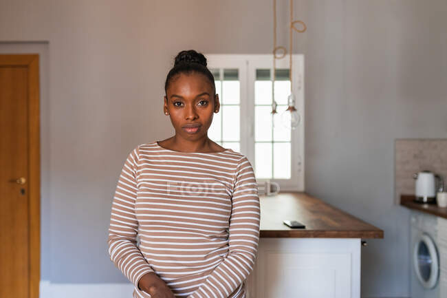 Молода афроамериканка з волосяною кулькою дивиться на камеру за столом і пральну машину на кухні. — стокове фото