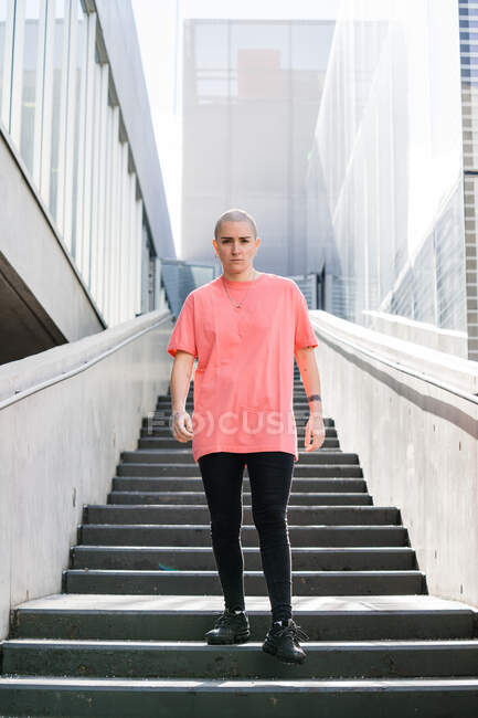 Transexuais pessoa em vestuário casual olhando para a câmera na escada da cidade entre casas contemporâneas à luz do sol — Fotografia de Stock