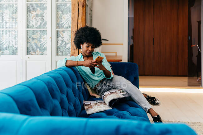 Belle jeune femme afro-américaine en vêtements décontractés naviguant sur smartphone et en lisant le magazine tout en se reposant sur un canapé bleu confortable à la maison — Photo de stock