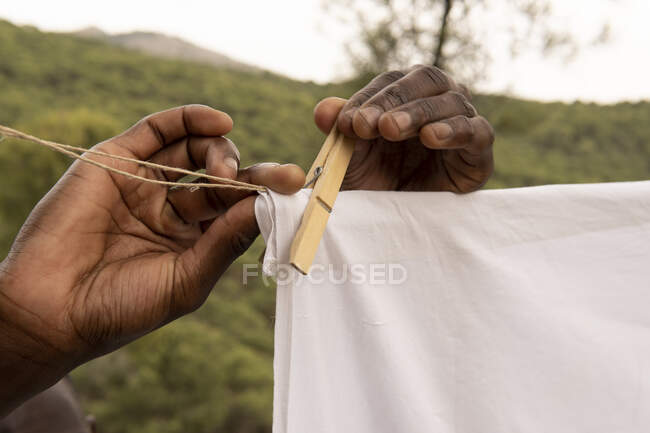 Cropped unrecognizable anonymous African American male hanging white cotton cloth on clothesline in countryside in summer — Stock Photo