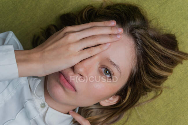 Visão superior da calma jovem fêmea de cabelos longos com olhos verdes cobrindo metade do rosto com a mão e olhando para a câmera — Fotografia de Stock