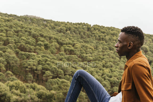 Vista lateral del pensativo hombre afroamericano en ropa de moda sentado en la naturaleza y admirando la vista del bosque montañoso - foto de stock
