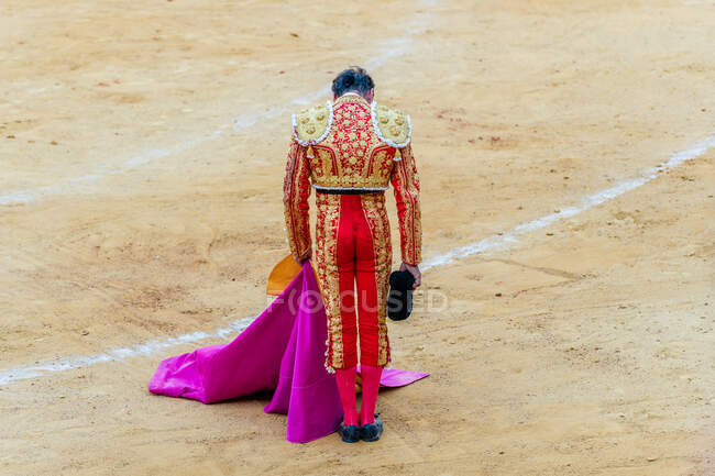 Back view of unrecognizable bullfighter in fancy costume taking off hat after corrida performance while standing on sandy arena — Stock Photo