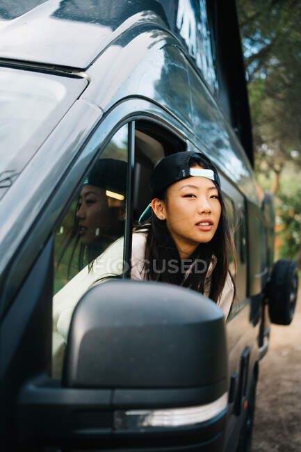 Glückliche junge asiatische Reisende mit Mütze, die aus dem offenen Fenster ihres Wohnmobils blickt und die Freiheit während ihrer Reise durch die sommerliche Landschaft genießt — Stockfoto