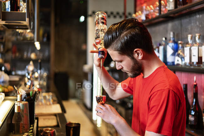 Вид збоку концентрованого бармена, що вливає алкоголь з пляшки в металевий джиґер під час приготування коктейлю в барі — стокове фото
