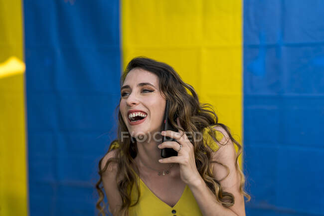 Positive female with wavy hair having conversation on mobile phone while laughing on two colored background in street and looking away — Stock Photo