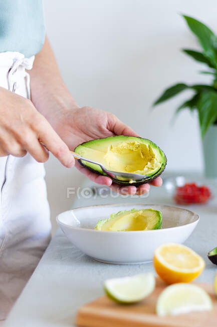 Colheita fêmea irreconhecível extraindo polpa de abacate madura com colher enquanto prepara guacamole à mesa com frutas cítricas em casa — Fotografia de Stock