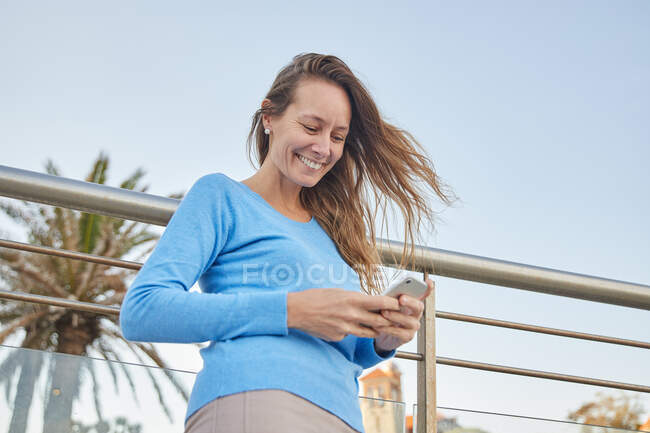 D'en bas de dame adulte souriante en vêtements décontractés debout près de la clôture et du vieil immeuble tout en surfant sur le téléphone dans le quartier de la ville en plein jour — Photo de stock