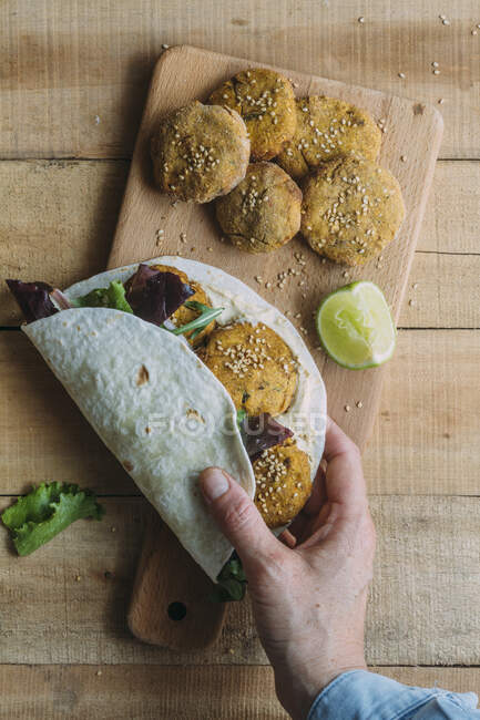 Top view of anonymous person wrapping fresh sweet potato falafel and lettuce into tortilla over cutting board on lumber table — Stock Photo