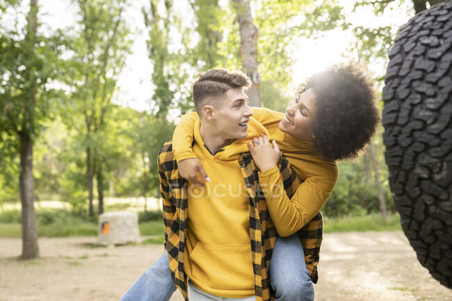 Junger Mann gibt optimistischen schwarzen Freundin Freizeitkleidung huckepack, während er in der Nähe von Auto an einem sonnigen Tag auf dem Land steht — Stockfoto