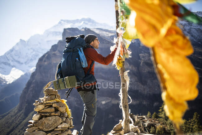 Vista lateral del caminante masculino colgando la bandera de oración budista en la cuerda durante el trekking en las montañas del Himalaya en Nepal - foto de stock