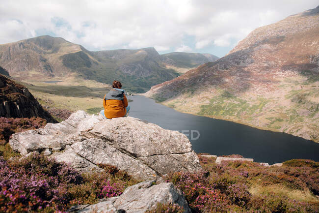 Задний вид анонимного мужчины, любующегося холмами и рекой, сидящего на грубом склоне во время путешествия по Сноудонии, сельская местность Великобритании — стоковое фото